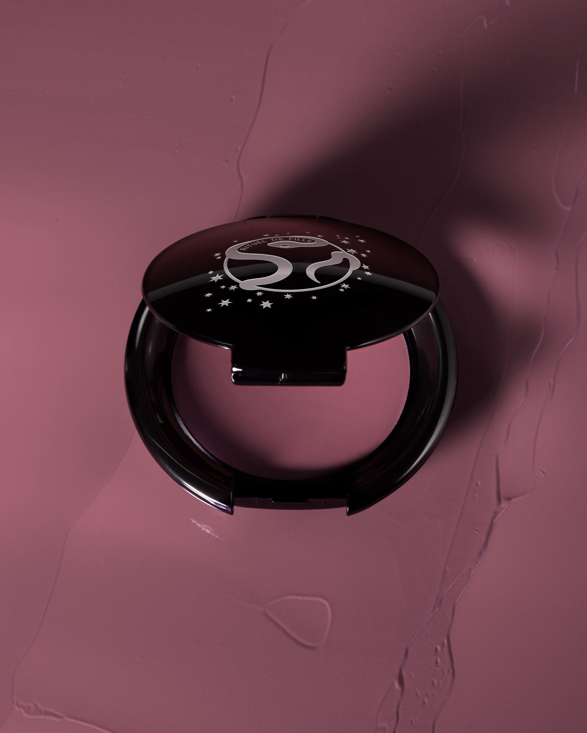 A round black makeup compact with a glossy lid featuring a white stylized logo and stars, set against a textured mauve-pink background with a matching makeup smear.